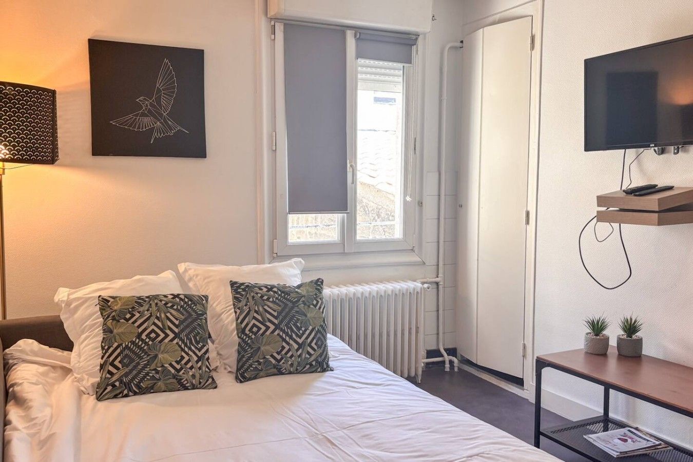 A bedroom with a bed, pillows, a radiator, a window, a television, and a small table with plants.