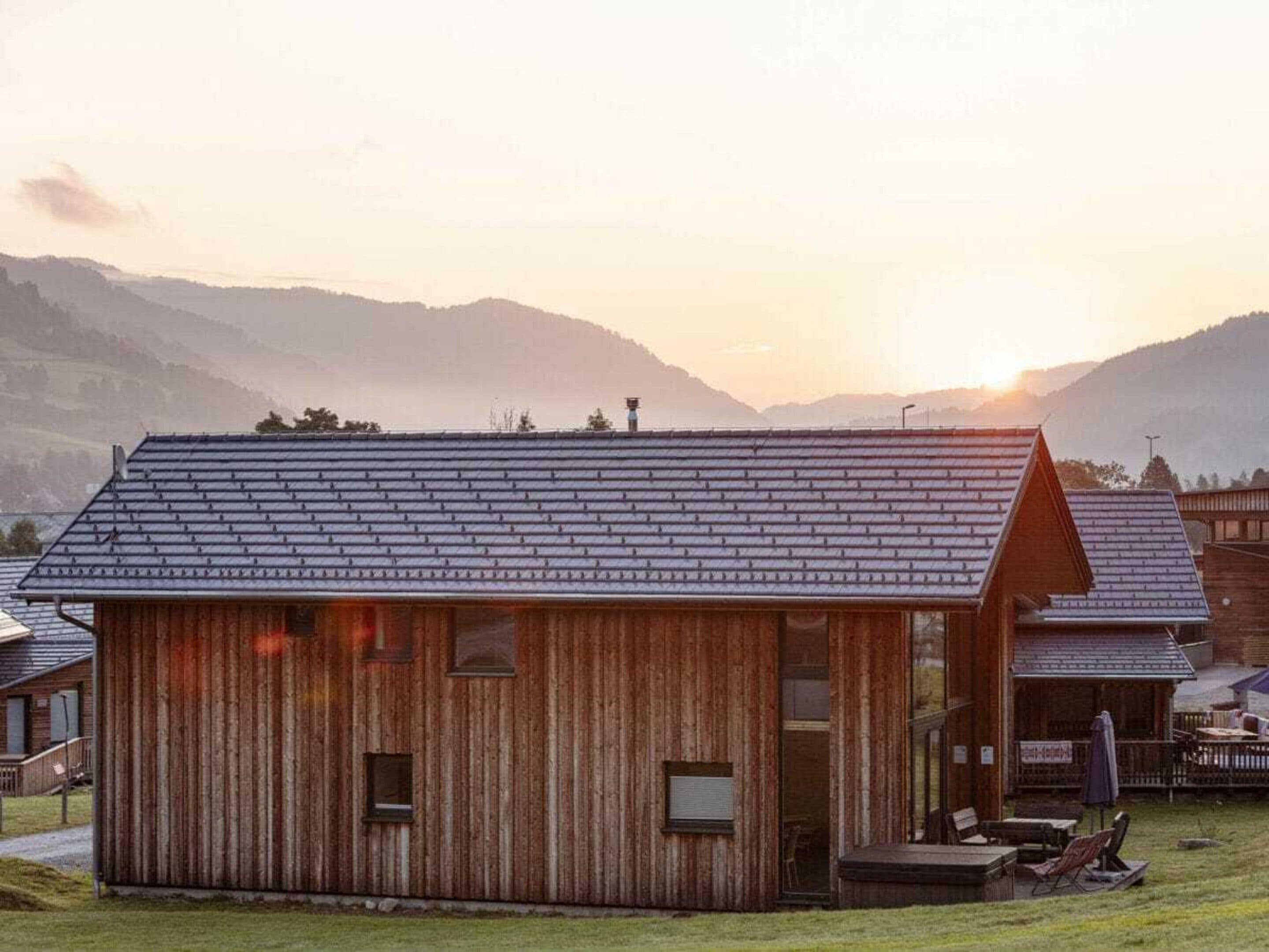 A rustic wooden cabin with a sloped roof, a porch with seating, and a view of the mountains in the background.