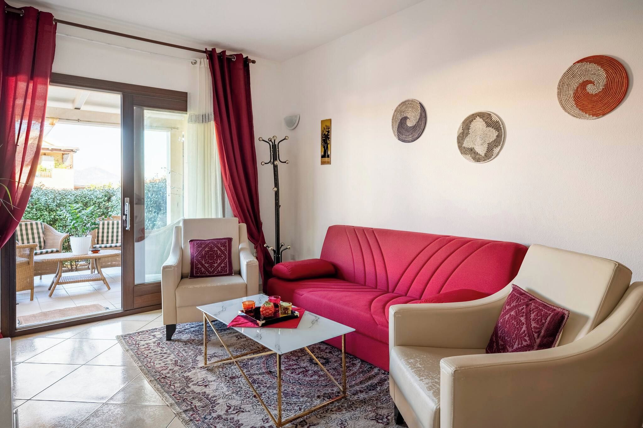 Ein Wohnzimmer mit einem roten Sofa, beigen Sesseln, einem Glastisch und einem gemusterten Teppich.