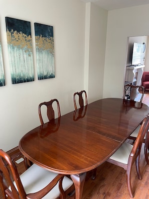 A dining room with a wooden table and chairs, two abstract paintings on the wall, and a television in the corner.