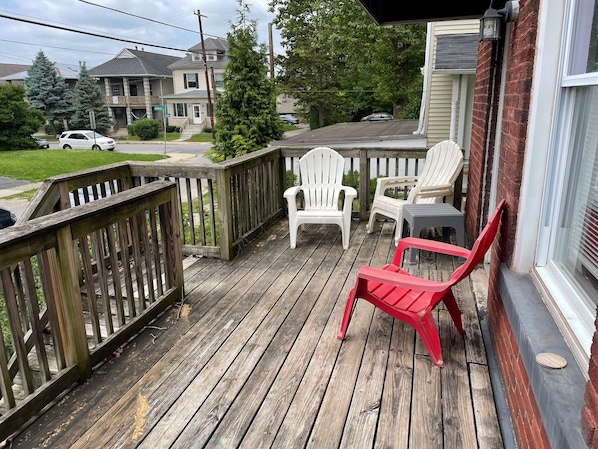 A wooden deck with two white chairs and one red chair, overlooking a street with houses and cars.