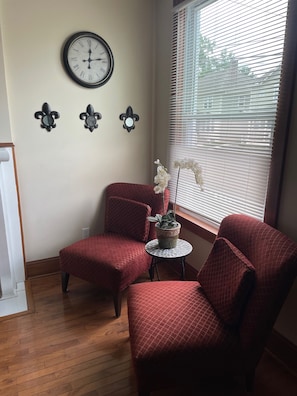 A room with two red upholstered chairs, a small round table, a wall clock, and a window with blinds.