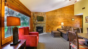 Ein gemütliches Wohnzimmer mit Kamin aus Stein, Holzwänden, einem roten Sessel, einem braunen Sofa und einem Esstisch mit Stühlen.