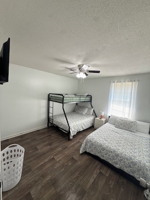 Quarto com cama beliche, uma cama de solteiro, ventilador de teto, janela com cortinas e TV fixada na parede.
