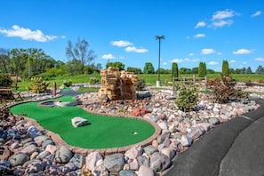 Un campo de mini golf con una superficie de césped verde, una formación rocosa como obstáculo y una valla de madera al fondo.