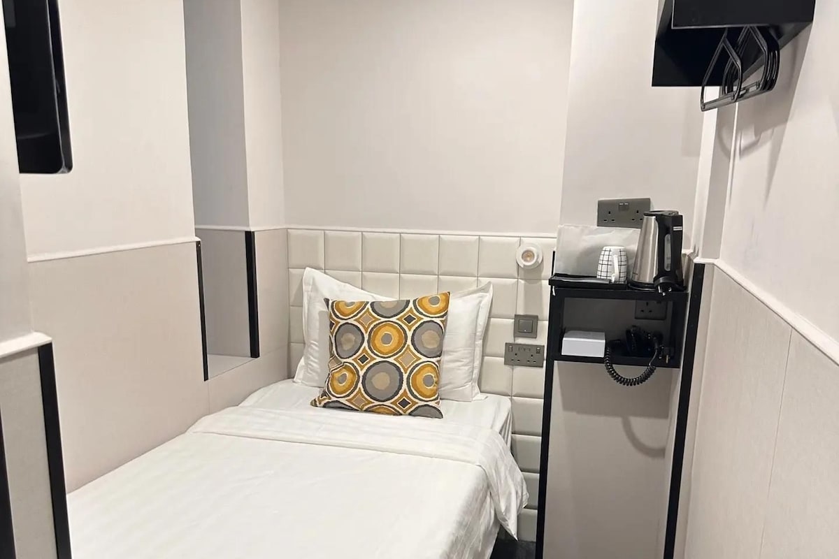 Una habitación de hotel pequeña y funcional con una cama individual, una mesita de noche con teléfono y un televisor montado en la pared.