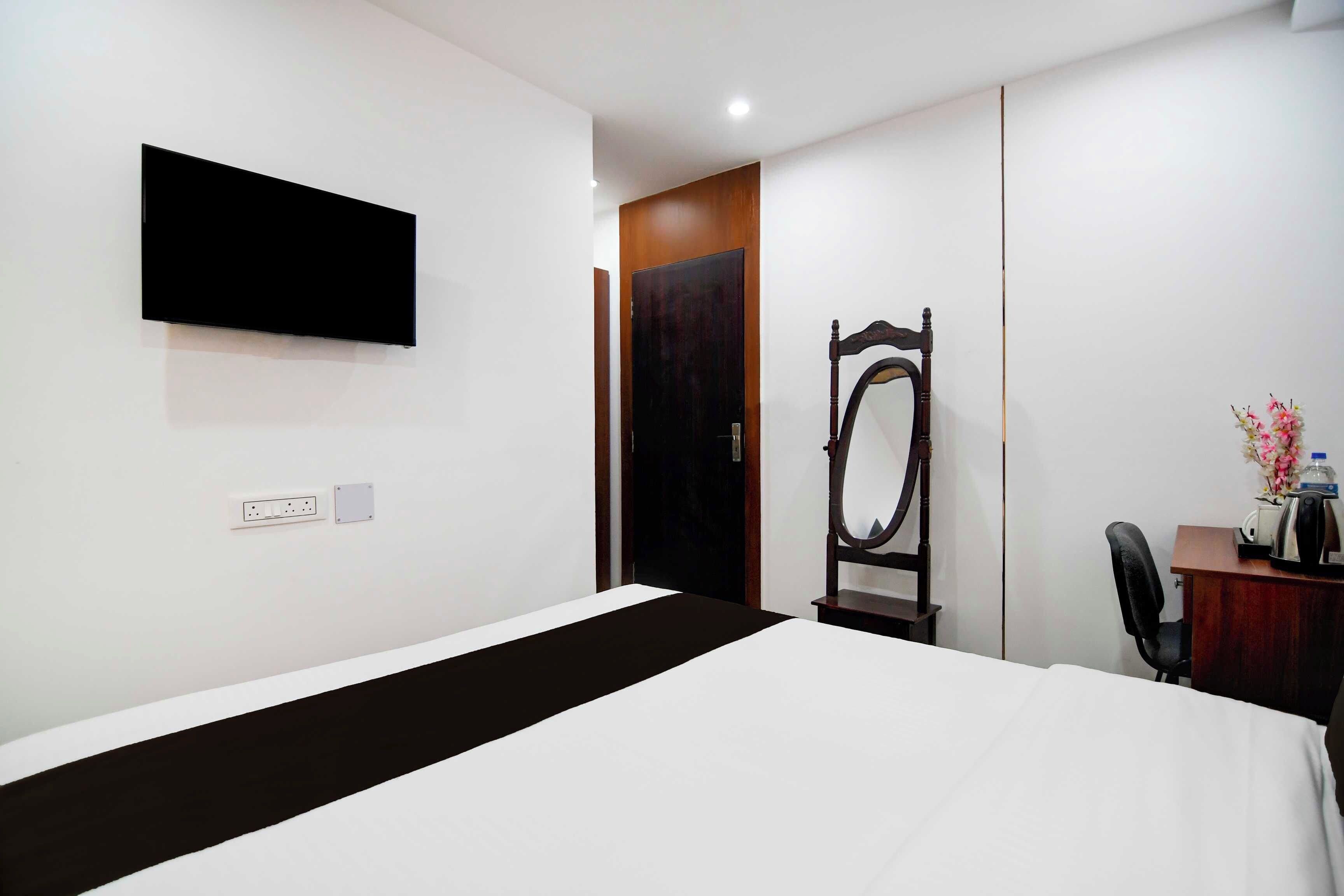 Habitación de hotel con una cama, un televisor de pantalla plana montado en la pared, una puerta de madera, un espejo y un escritorio con una tetera y flores.