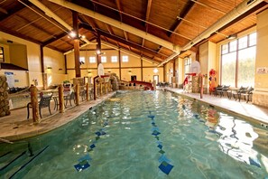 An indoor swimming pool with a wooden ceiling, a diving board, and a seating area with tables and chairs. - Wisconsin Dells | Waterpark Launchpad | Pools + Golf | 2BR (Wisconsin Dells)