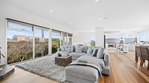 A modern living room with a grey sofa, a coffee table, and a view of a kitchen and outdoor scenery through large windows. - Saltaire - stunning 4 bedroom property with indoor pool (St Andrews Beach)
