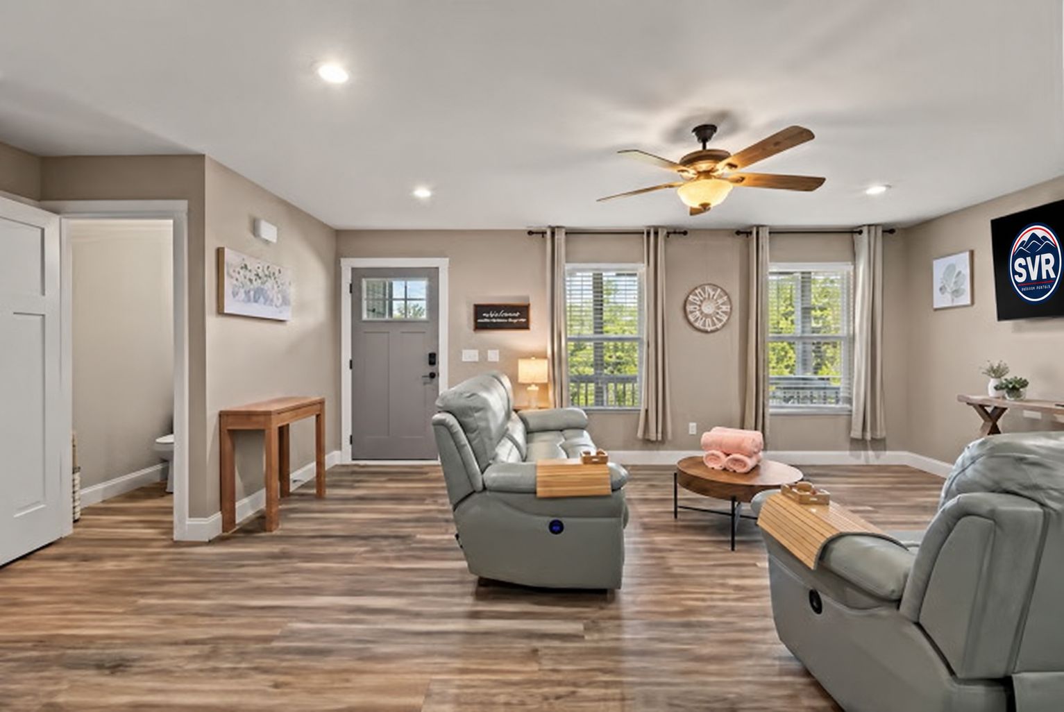 A spacious living room with a ceiling fan, a grey leather sofa, a wooden coffee table, and a door leading to another room.