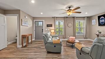 A spacious living room with a ceiling fan, a grey leather sofa, a wooden coffee table, and a door leading to another room.