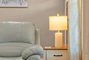 A room with a light-colored sofa, a bedside table with a lamp, and a wall-mounted artwork.