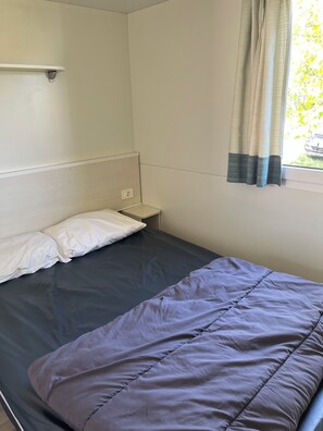 Ein Einzelbett mit dunkelbrauner Matratze und hellem Kopfkissen, ein kleiner Nachttisch, ein an der Wand befestigter Lichtschalter und ein Fenster mit Vorhängen.
