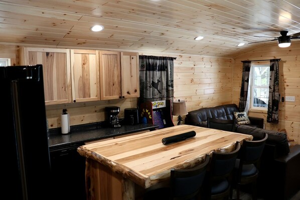 A wooden cabin interior with a kitchen area featuring wooden cabinets, a black refrigerator, a coffee maker, and a roll of paper towels. There is a dining table with chairs and a black leather sofa with a pillow.