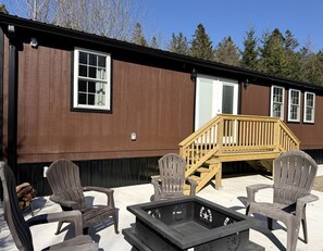 A wooden deck with Adirondack chairs and a fire pit, adjacent to a brown building with windows and a door.