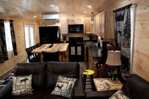 A cabin interior with a kitchenette, a sofa with decorative pillows, a small table with a lamp, and a window with curtains.