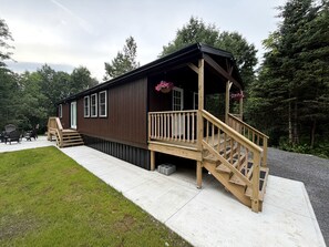 A wooden cabin with a porch and stairs, surrounded by a grassy area and trees.