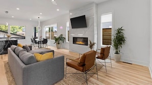 Een moderne woonkamer met een open haard, een grijze bank, een bruine lederen fauteuil en een eethoek met een tafel en stoelen.