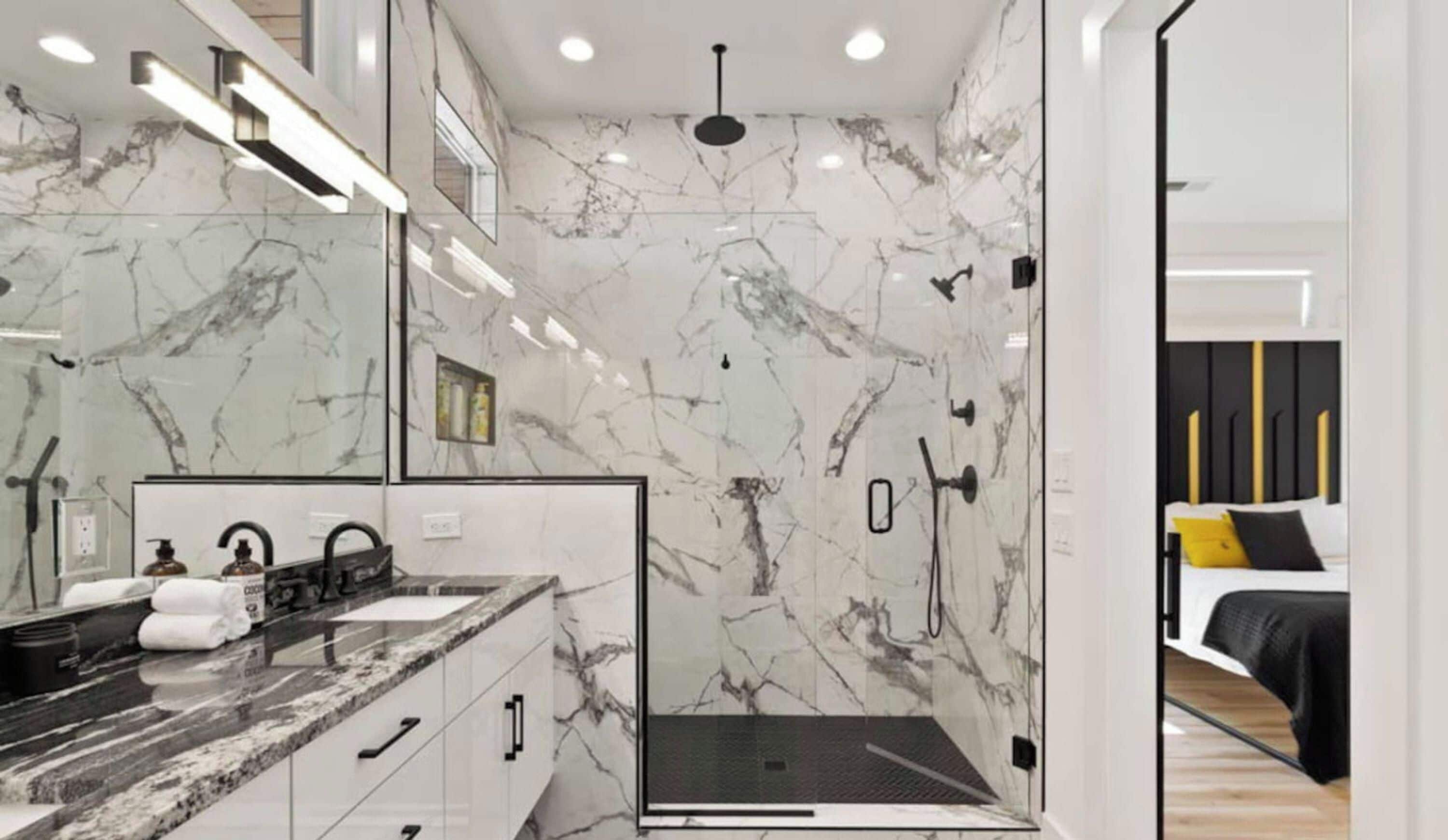 A modern bathroom with a marble wall, a glass shower enclosure, and a visible bed with black and yellow bedding in the adjacent room.