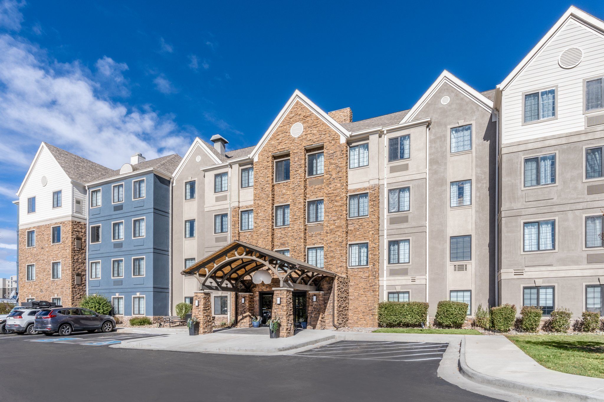 A multi-story hotel with a central entrance, parking area, and a clear blue sky.
