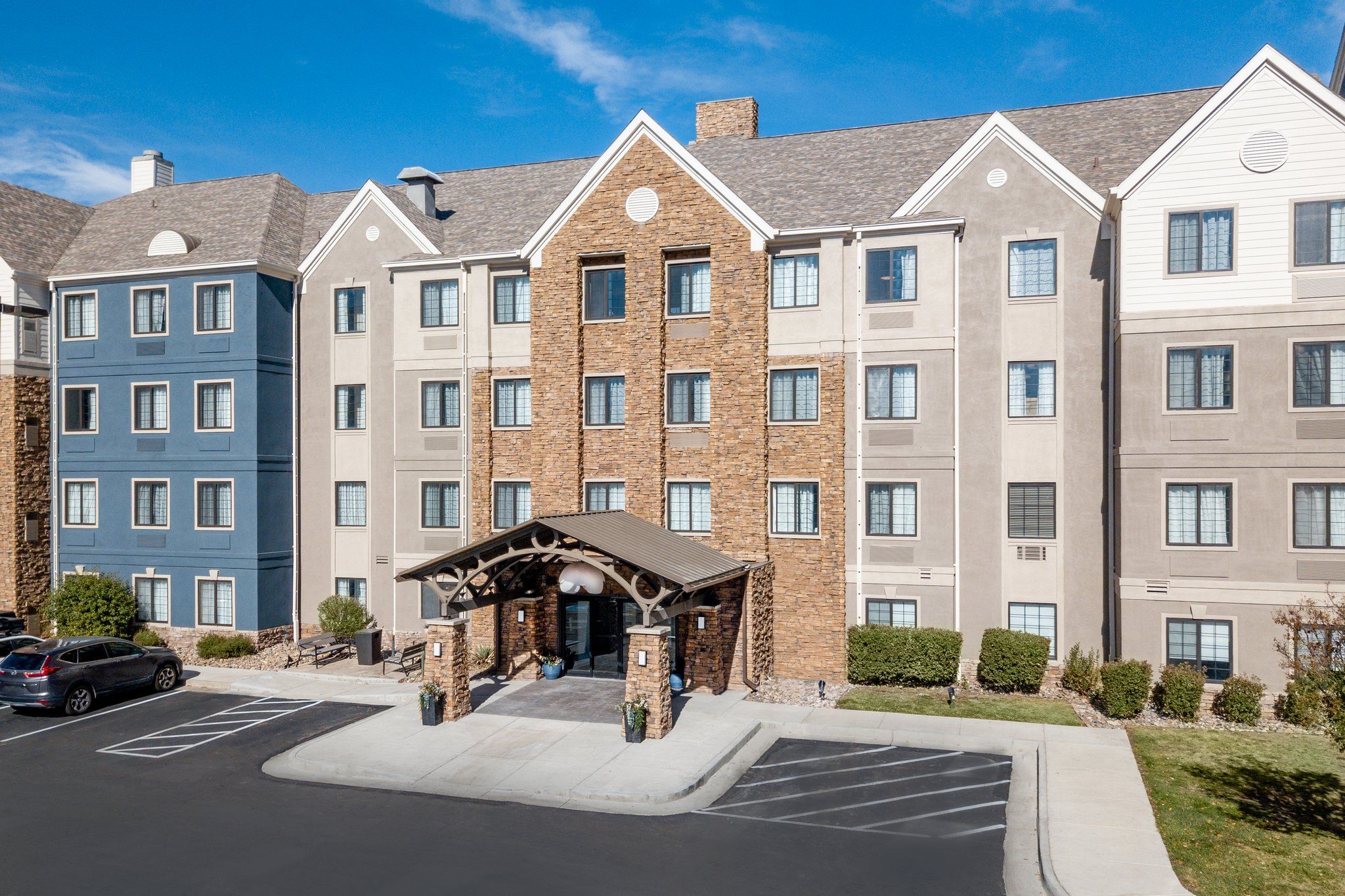 A multi-story hotel with a central entrance, parking lot, and a clear blue sky.