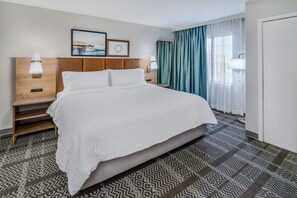 Quarto de hotel com uma cama grande, duas mesas de cabeceira com abajures, cabeceira, quadro emoldurado e janela com cortinas.