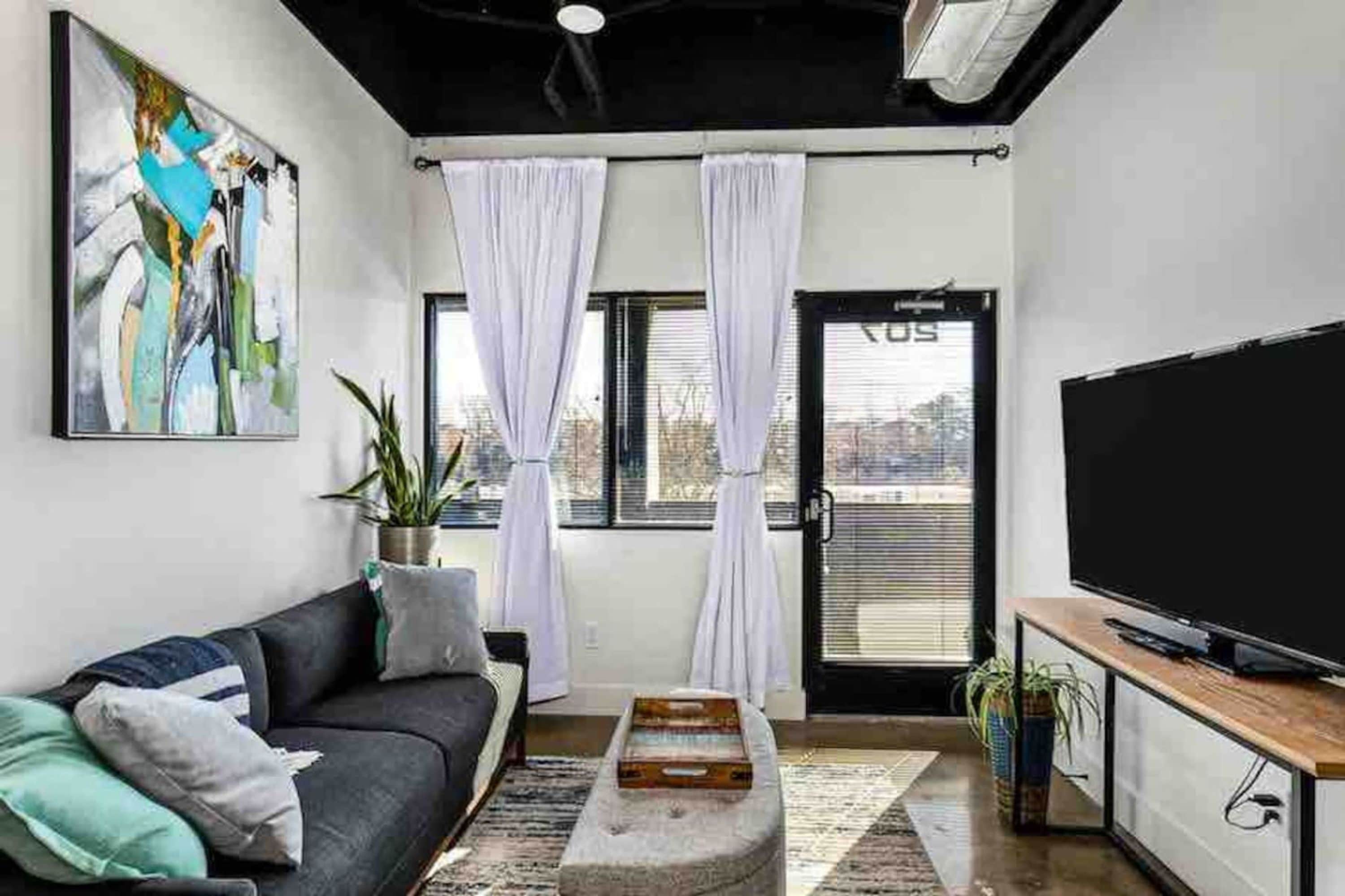 A modern living room with a black sofa, a flat-screen TV, and a large window with white curtains.