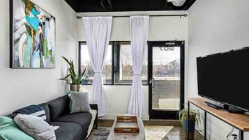 A modern living room with a black sofa, a flat-screen TV, and a large window with white curtains.