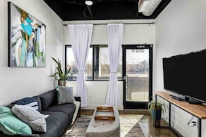 A modern living room with a black sofa, a flat-screen TV, and a large window with white curtains. - Condo Minutes to Downtown - pet Friendly w Parking (Nashville)