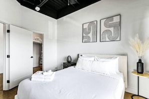 A modern bedroom with a large bed, a bedside table, a nightstand, and a small round table with a vase. There is a closet with a hanging rack and a door leading to another room. - Condo Minutes to Downtown - pet Friendly w Parking (Nashville)