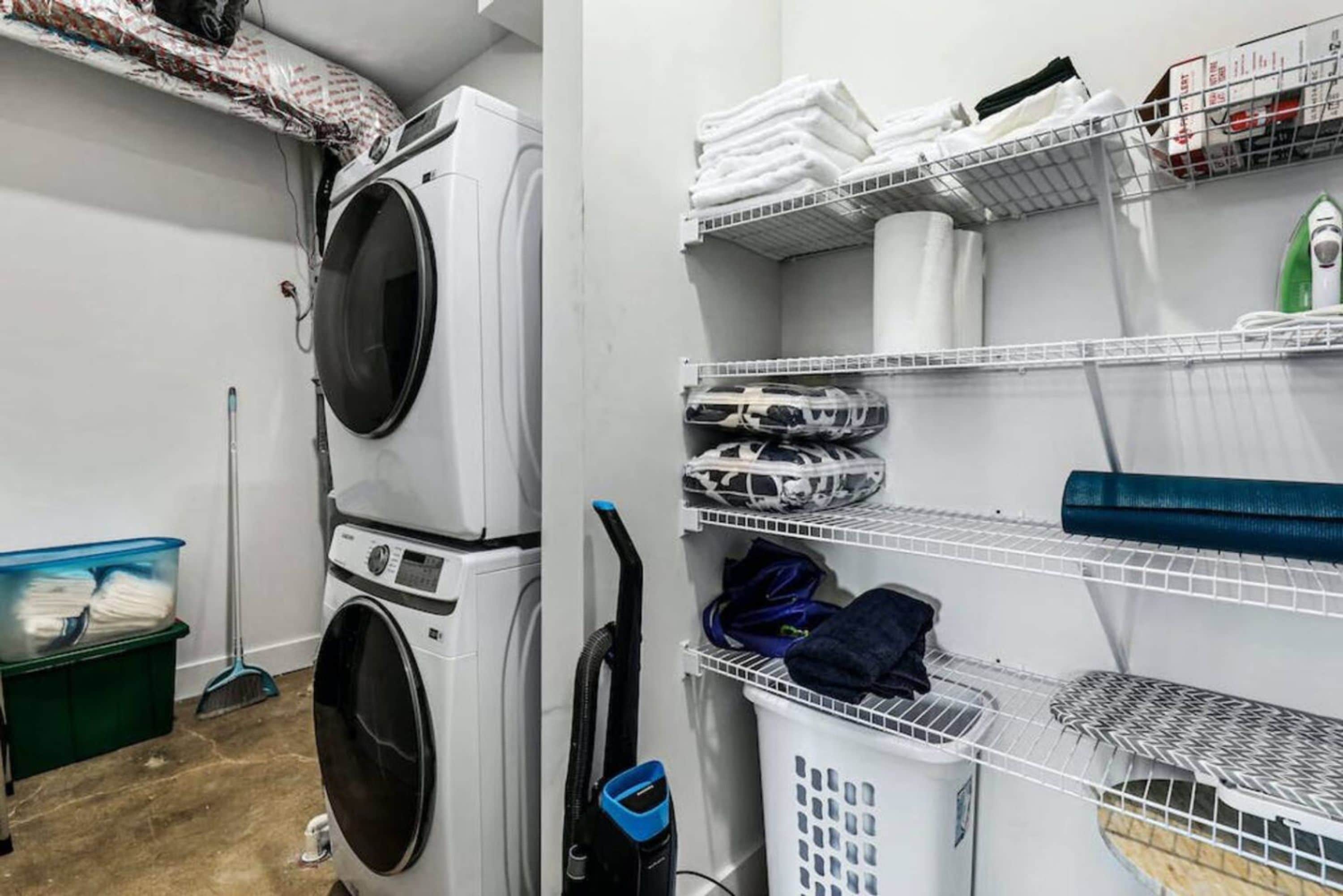 A laundry room with stacked washing machines, shelves with folded towels, and a green laundry basket.