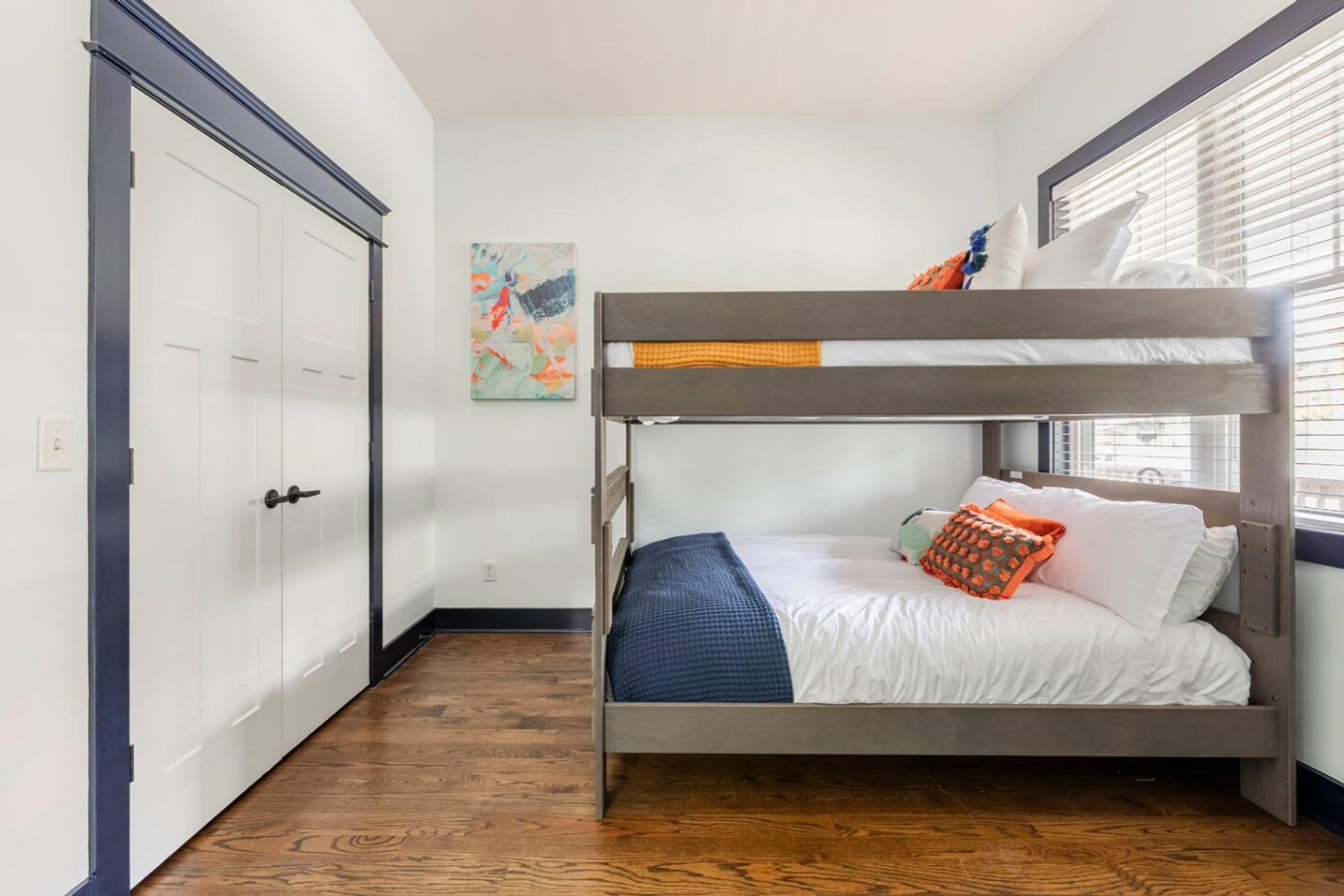 Ein Etagenbett mit weißer Matratze und blauer Decke, ein Holzboden, eine weiße Tür und ein Fenster mit Jalousien.