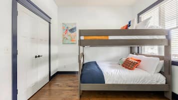A bunk bed with a white mattress and blue blanket, a wooden floor, a white door, and a window with blinds.