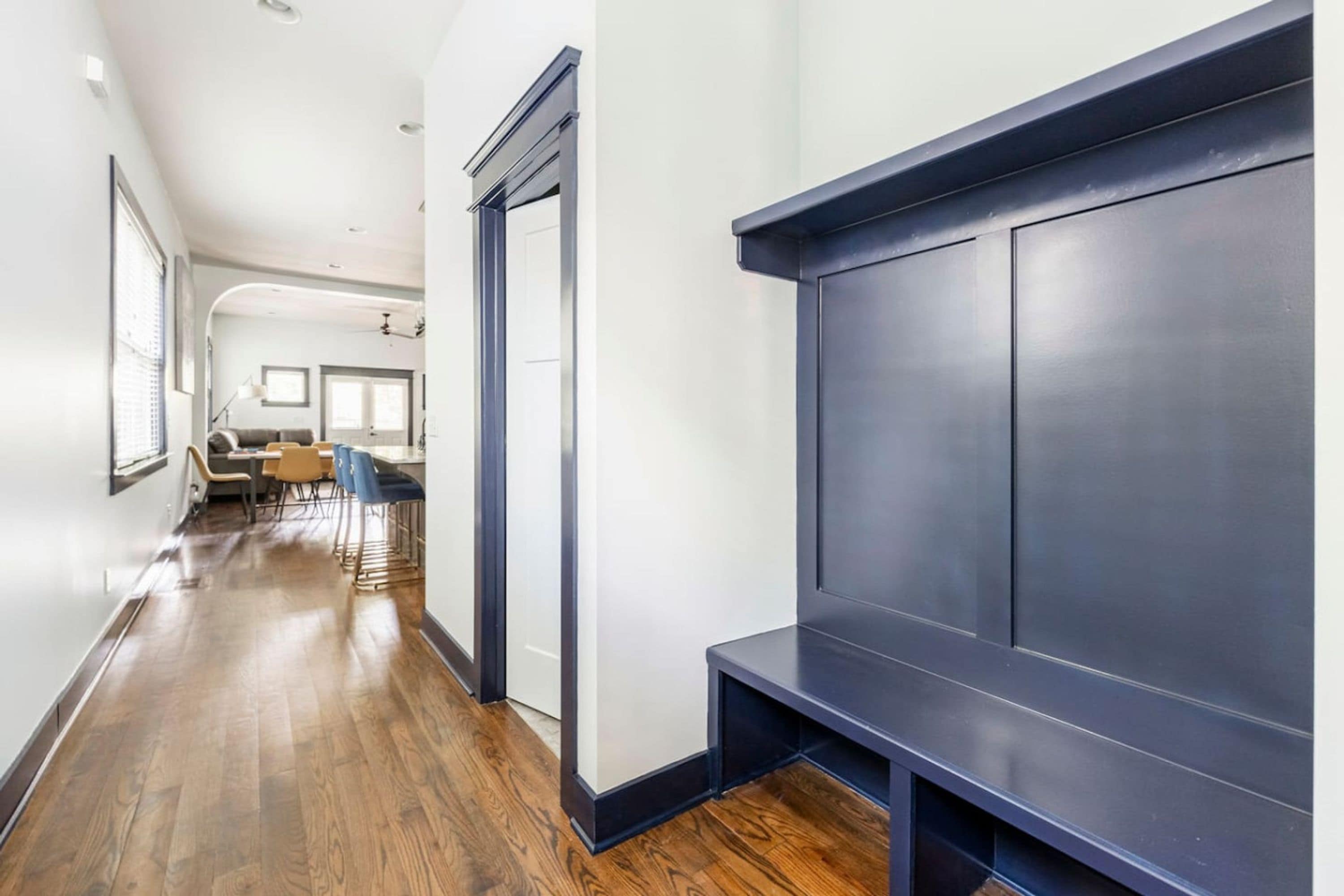 A hallway with a dark blue accent wall, wooden flooring, and a door leading to a dining area with chairs and a table.