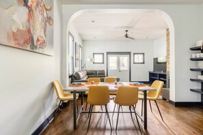 Zona de dinar amb taula preparada per a quatre persones, envoltada de cadires grogues, a una habitació amb terra de fusta i un ventilador de sostre.
