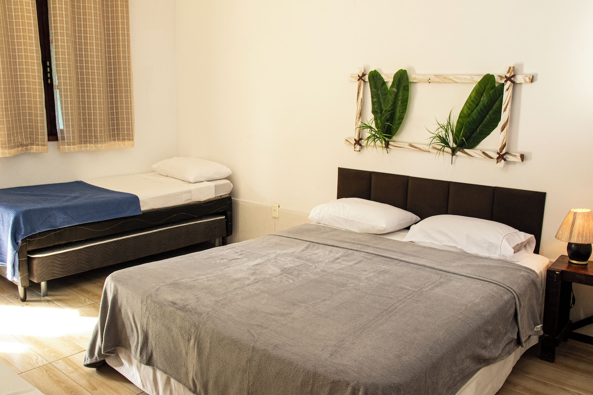 A bedroom with two beds, a nightstand with a lamp, a window with curtains, and a wall-mounted plant decoration.