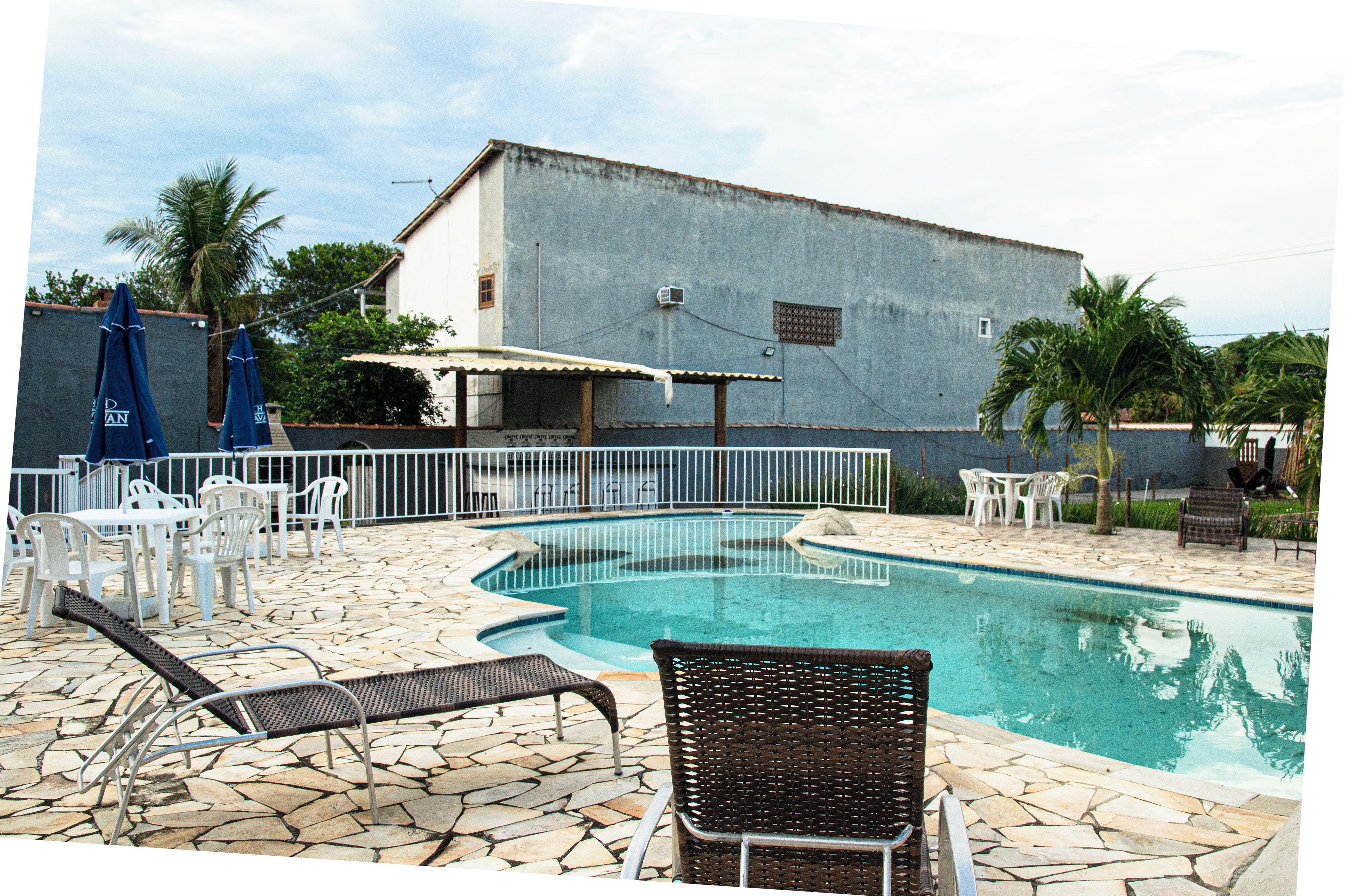 A swimming pool with lounge chairs and tables, surrounded by a stone patio and a building in the background.