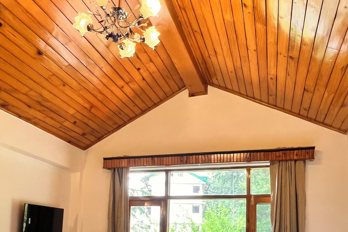 Quarto de hotel com teto de madeira, cama com roupa de cama verde e branca, escrivaninha com chaleira, televisão e janela com cortinas.