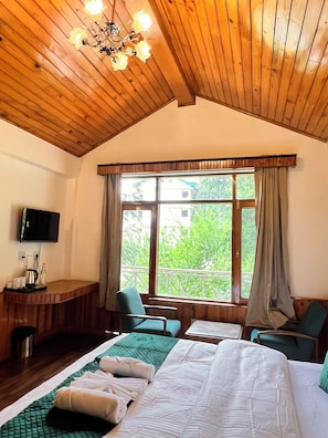 Quarto de hotel com teto de madeira, cama com roupa de cama verde e branca, escrivaninha com chaleira, televisão e janela com cortinas.