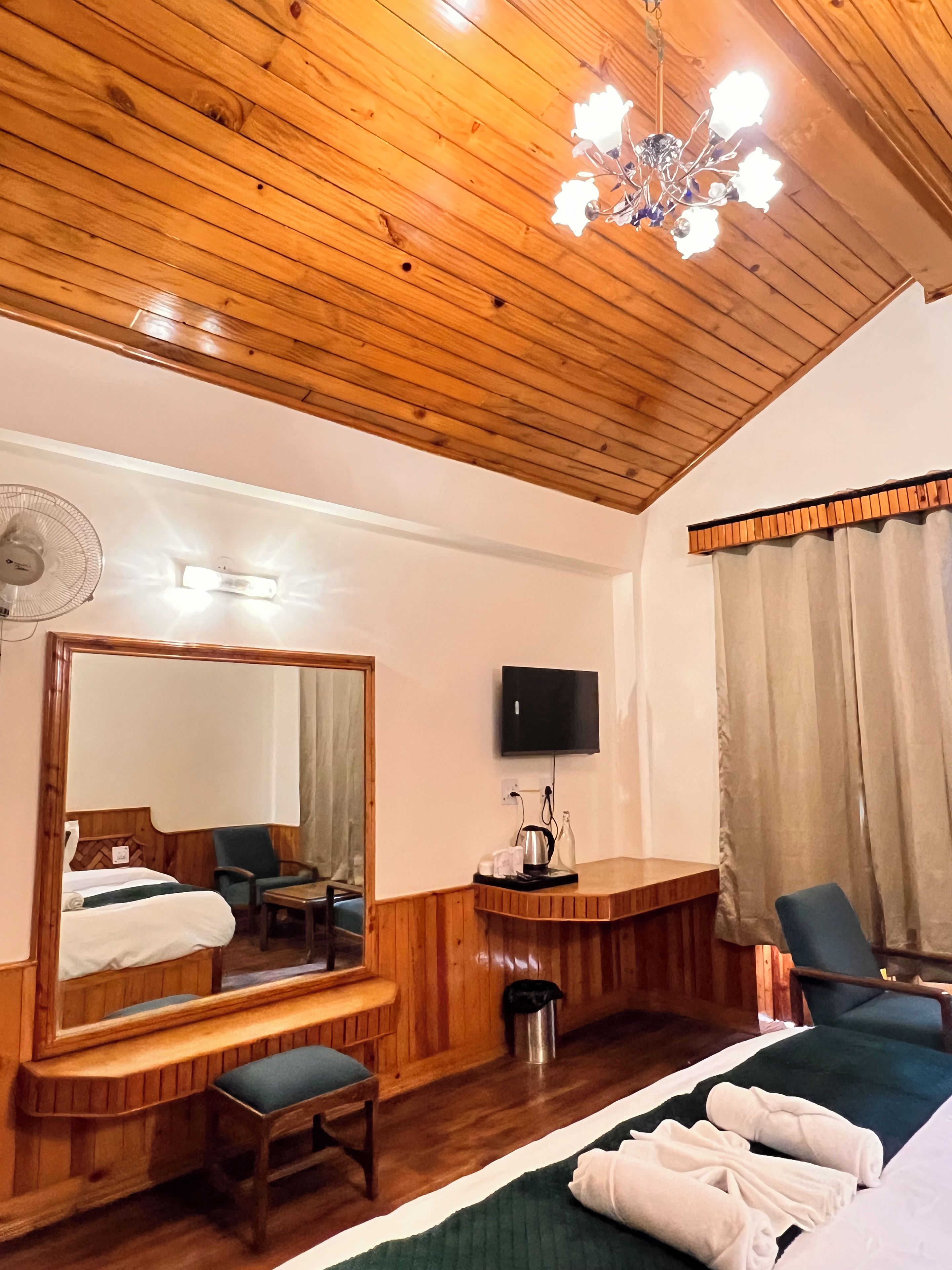 Una camera d'albergo con soffitto in legno, un grande specchio, un letto con lenzuola bianche, una scrivania con televisore e una sedia.