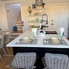 Un comedor con una mesa blanca dispuesta para cuatro, una cocina con un grifo y una escalera que lleva a otra habitación.