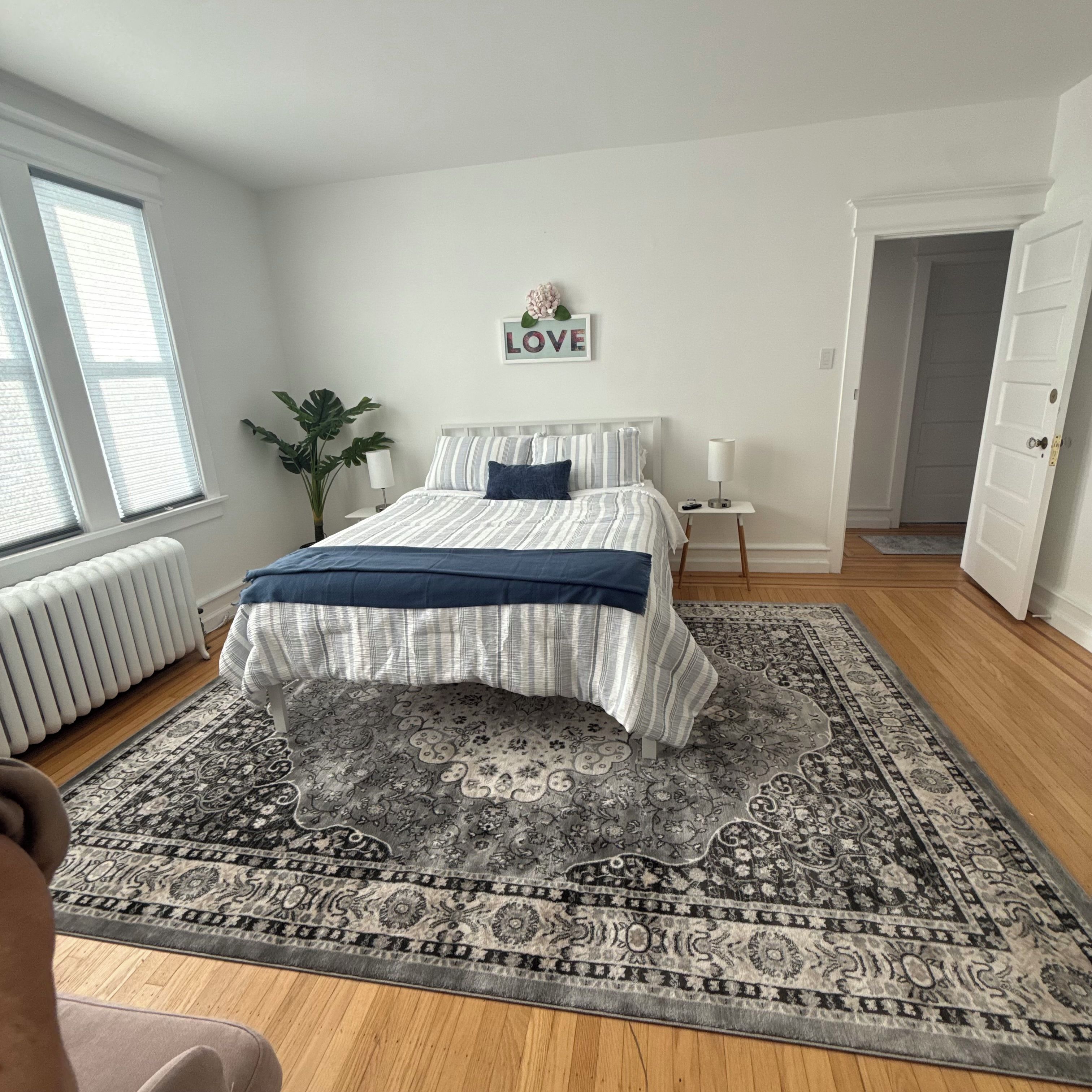 Ein Schlafzimmer mit einem Bett, einem Heizkörper, einem großen Teppich, einer Pflanze und einem 'LOVE'-Schild an der Wand.