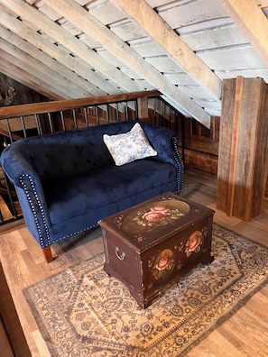 A blue sofa with a floral pillow, a wooden chest of drawers with floral designs, and a patterned rug in a room with wooden flooring and a sloped ceiling.