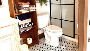 A bathroom with a toilet, a shower enclosure, a wooden shelf with towels, and a patterned floor. - Adorable Old World Livery Stable with balcony overlooking Downtown Madison (Madison)