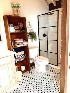 A bathroom with a toilet, a shower enclosure, a wooden shelf with towels, and a patterned floor. - Adorable Old World Livery Stable with balcony overlooking Downtown Madison (Madison)