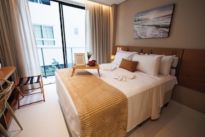 Quarto de hotel com cama, mesa de cabeceira com abajur, uma cadeira e janela com cortinas.