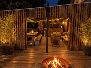 En udendørs træterrasse med bålsted, siddepladser og lysslynge.
