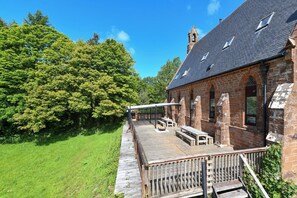 En murstensbygning med en træterrasse, udendørs siddepladser og et stort træ i baggrunden.