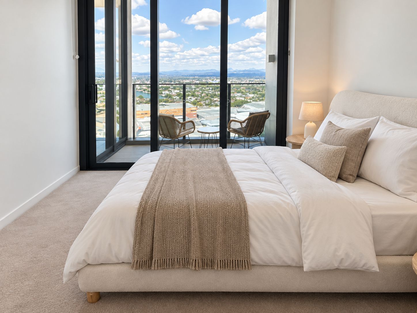 A bedroom with a large bed, a view of a balcony with outdoor furniture, and a cityscape in the background.