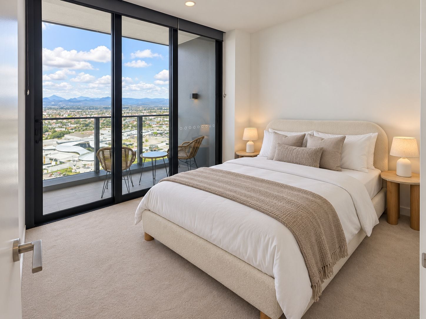 Ein Schlafzimmer mit einem großen Bett, zwei Nachttischlampen, Blick auf eine Stadtkulisse durch eine Schiebetür und einem Balkon mit Stühlen.